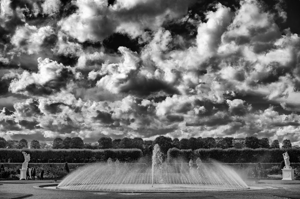 Schwarzweiß-Bild. Ein Fontänen-Brunnen mit Mittelstrahl. Ringförmig von außen spritzen weitere Düsen Wasser zur Mitte.
Wolkiger Himmel mit starkem Kontrast